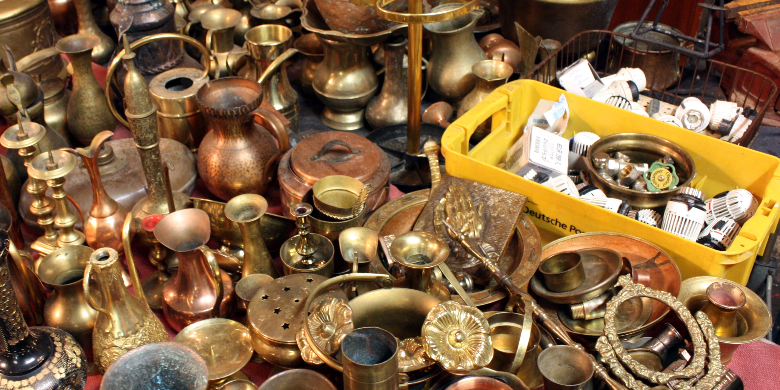 Foto auf einem Trödelmarkt: viele messing- und kupferfarbene alte Vasen, Töpfe und andere Behälter sind von schräg oben zu sehen. Dazwischen ein Messing-Relief zweier betender Hände. Auf der rechten Seite steht eine gelbe Kunststoffkiste der deutschen Post mit Kleinteilen (z.B. Heizungsthermostate).